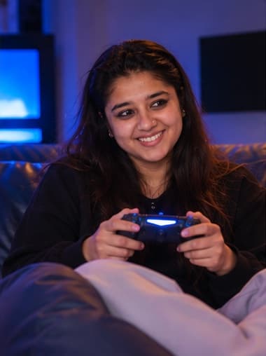 Editorial artwork: Abhilasha holding a game controller in a dim room lit by a screen.