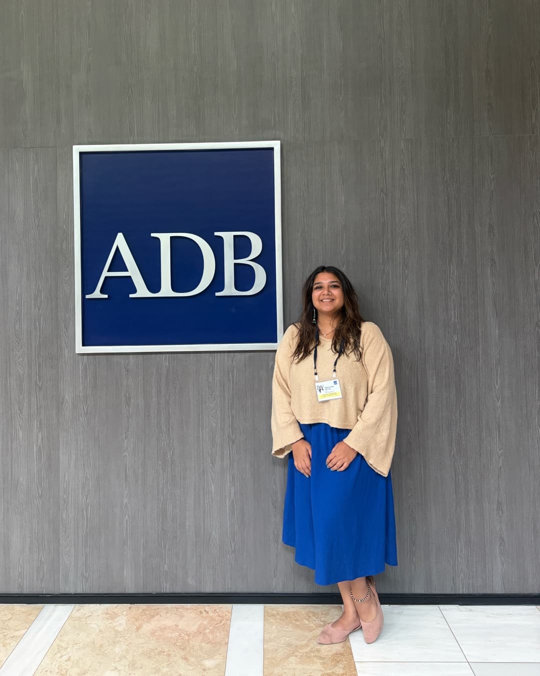 Abhilasha Mittal smiling beside the ADB sign, in a beige jumper and a blue skirt.